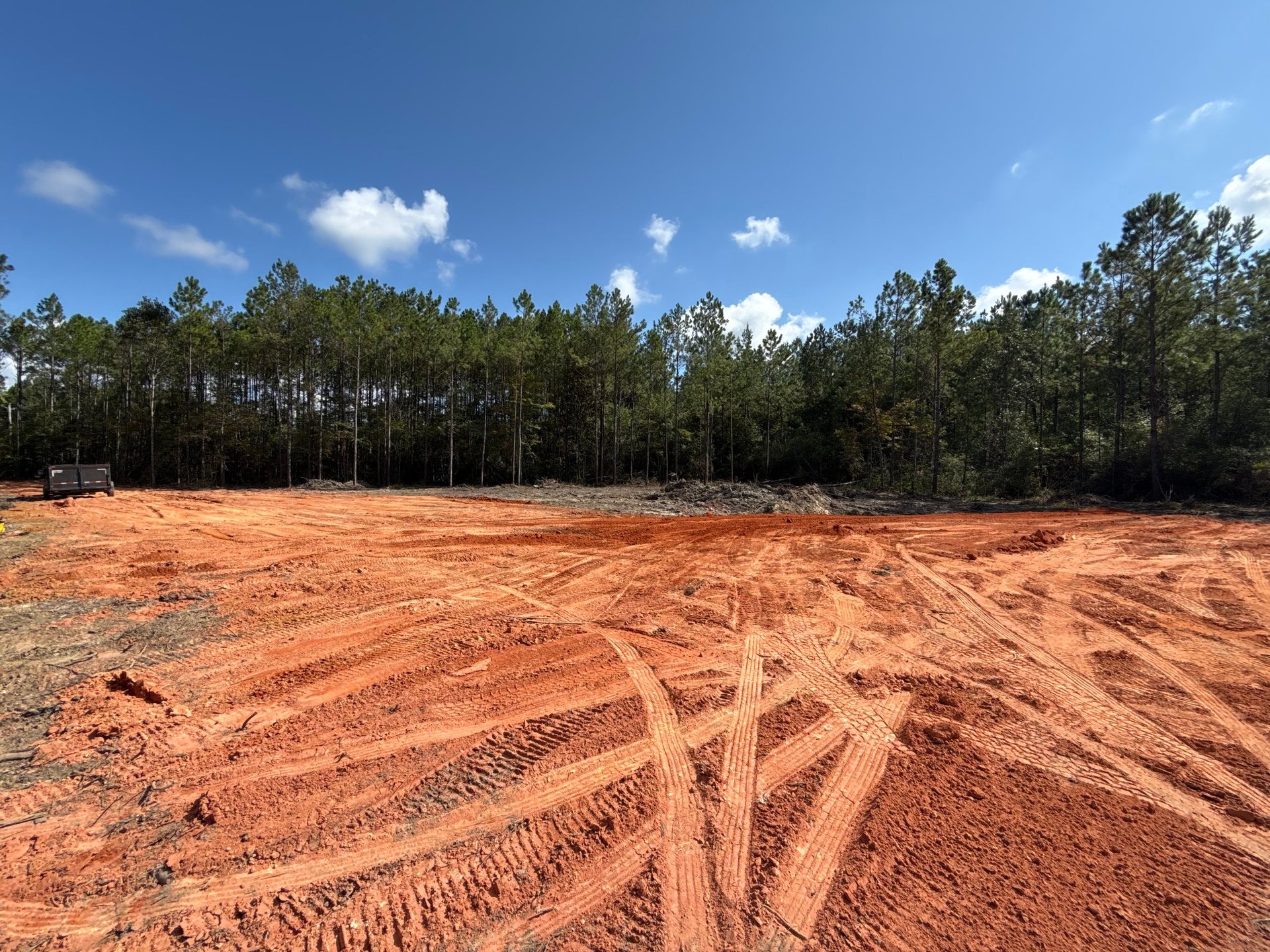 Cleared red dirt construction site with tire tracks and forest treeline under blue sky